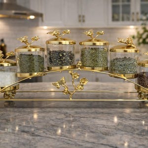 Puede incluir: Un especiero dorado con seis tarros de vidrio transparente llenos de especias. Cada tarro tiene una tapa dorada con un diseño floral. El especiero presenta un diseño arqueado con detalles florales.