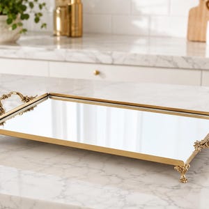 Puede incluir: Una bandeja rectangular con espejo y marco de metal en tono dorado pulido. La bandeja incluye asas decorativas ornamentadas en ambos lados y se apoya sobre cuatro pequeñas patas de metal intrincadas en cada esquina, creando una base elevada.