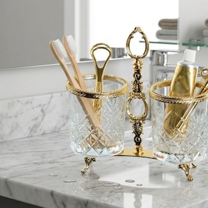 May include: A decorative bathroom organizer with two clear glass holders, each with a gold rim and ornate gold feet. One holder contains toothbrushes and a tongue scraper, the other a tube of toothpaste and a key. The stand is gold.