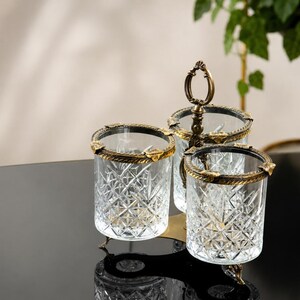 May include: A decorative glass serving set with three compartments. Each glass has a diamond pattern and is trimmed with gold-colored metal. The set includes a handle for easy carrying and is placed on a black surface.