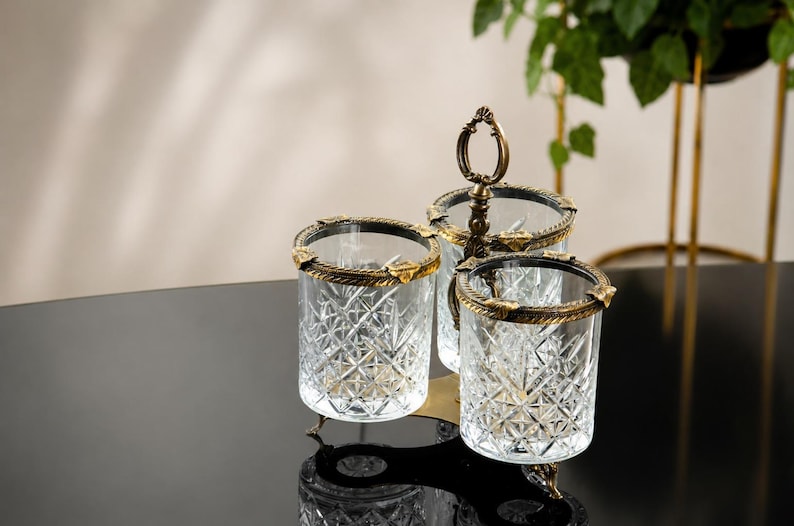 May include: A decorative glass serving set with three compartments. Each glass has a diamond pattern and is trimmed with gold-colored metal. The set includes a handle for easy carrying and is placed on a black surface.