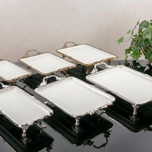 Könnte beinhalten: Sechs rechteckige Serviertabletts mit weißen Oberflächen und verzierten Metallrahmen. Die Tabletts sind unterschiedlich groß und haben dekorative Griffe. Die Metallrahmen sind entweder silber- oder goldfarben. Eine dekorative Pflanze in einem bronzefarbenen Topf befindet sich im Hintergrund.