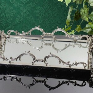Pu&ograve; includere: Un vassoio rettangolare argentato con dettagli ornati e decorazioni a volute. Il vassoio ha una superficie a specchio e manici decorativi. Il design presenta curve e decorazioni simmetriche, che riflettono uno stile classico ed elegante. Il vassoio &egrave; probabilmente utilizzato per servire o esporre.
