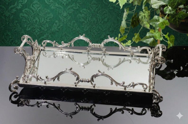 Pu&ograve; includere: Un vassoio rettangolare argentato con dettagli ornati e decorazioni a volute. Il vassoio ha una superficie a specchio e manici decorativi. Il design presenta curve e decorazioni simmetriche, che riflettono uno stile classico ed elegante. Il vassoio &egrave; probabilmente utilizzato per servire o esporre.