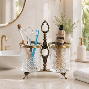 May include: A double-cup bathroom organizer featuring two clear, diamond-patterned glass tumblers held by an ornate, antique-bronze metal frame with a central handle and decorative feet, containing two toothbrushes, a tongue scraper, and a tube.