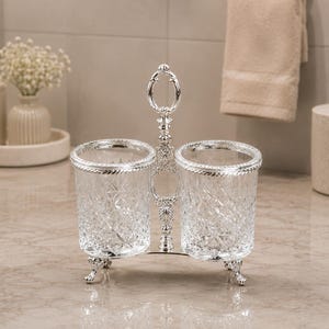 May include: A silver-toned, double-cup holder with ornate detailing. The clear glass cups have a diamond pattern and are trimmed with silver. The holder has a handle and rests on decorative feet. A bathroom accessory.