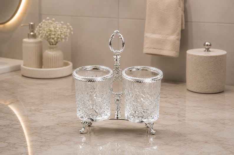 May include: A silver-toned, double-cup holder with ornate detailing. The clear glass cups have a diamond pattern and are trimmed with silver. The holder has a handle and rests on decorative feet. A bathroom accessory.