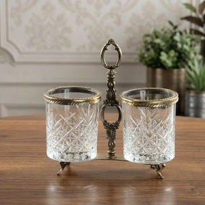 May include: A decorative glass and metal condiment holder with two clear glass containers. Each glass has a gold rim and sits on a decorative metal base. The holder has an ornate handle.