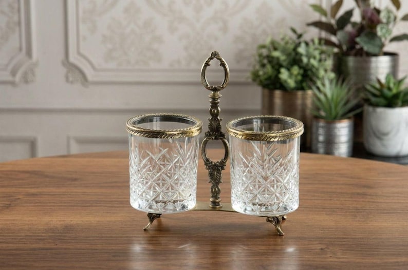 May include: A decorative glass and metal condiment holder with two clear glass containers. Each glass has a gold rim and sits on a decorative metal base. The holder has an ornate handle.