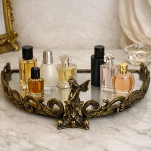 May include: A decorative vanity tray with a mirrored surface and ornate gold-colored filigree trim. The tray holds several perfume bottles of varying shapes and colors, and a small bronze fairy statue.