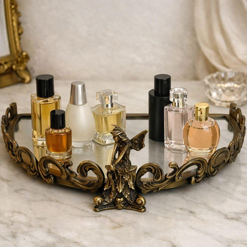 May include: A decorative vanity tray with a mirrored surface and ornate gold-colored filigree trim. The tray holds several perfume bottles of varying shapes and colors, and a small bronze fairy statue.