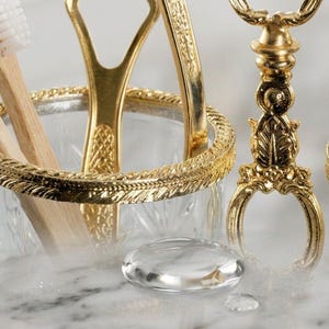 May include: Close-up of a gold-toned bathroom accessory set. The set includes a toothbrush holder with wooden toothbrushes, a tongue scraper, and a decorative stand. The items are set against a blurred background.