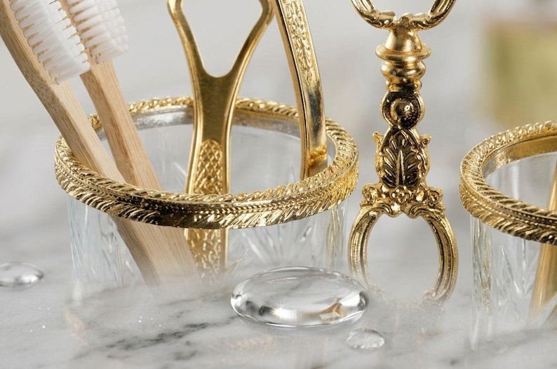 May include: Close-up of a gold-toned bathroom accessory set. The set includes a toothbrush holder with wooden toothbrushes, a tongue scraper, and a decorative stand. The items are set against a blurred background.