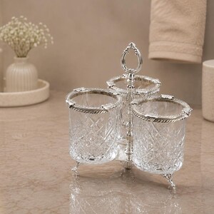 May include: A three-section crystal and silver-toned vanity organizer. Each section is a clear, cut-glass cup with a silver-toned rim and decorative legs. The organizer has a handle at the top. The background includes a soap dispenser, vase, and a small container.