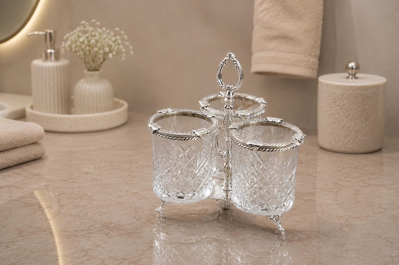 May include: A three-section crystal and silver-toned vanity organizer. Each section is a clear, cut-glass cup with a silver-toned rim and decorative legs. The organizer has a handle at the top. The background includes a soap dispenser, vase, and a small container.