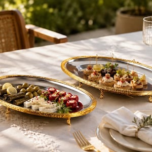 May include: Two oval serving trays with polished silver-tone centres and ornate, textured gold-tone rims, each supported by four small decorative feet. The trays are arranged on a table, holding various appetisers including olives and stuffed peppers.