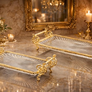 Könnte beinhalten: Zwei rechteckige Glastabletts mit goldfarbenen, verzierten Griffen und Verzierungen. Die Tabletts haben ein Sonnenstrahlenmuster auf dem Glas. Der Hintergrund umfasst Kerzen, Blumen und einen goldgerahmten Spiegel.