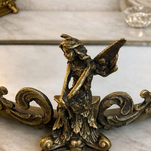 May include: A bronze-colored fairy figurine with detailed wings and a flowing dress. The fairy is positioned on an ornate, curved base with scrollwork. The sculpture is likely a decorative item.
