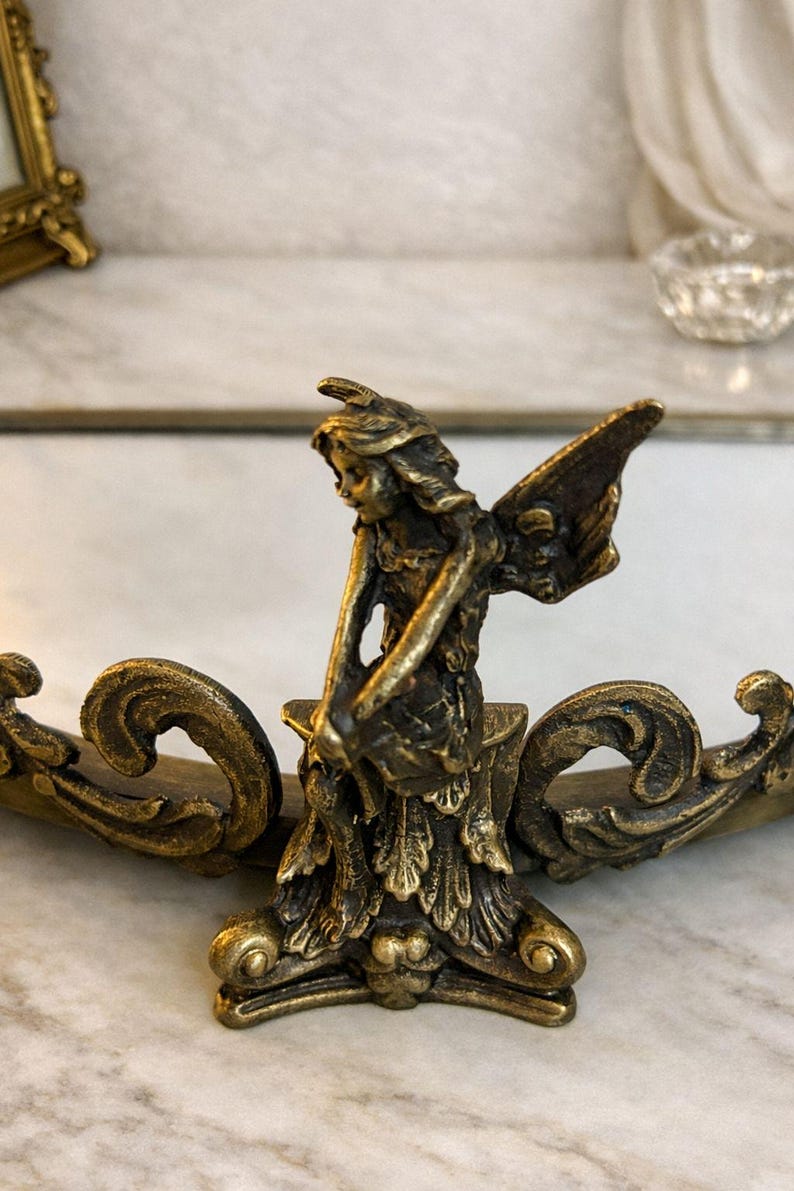 May include: A bronze-colored fairy figurine with detailed wings and a flowing dress. The fairy is positioned on an ornate, curved base with scrollwork. The sculpture is likely a decorative item.