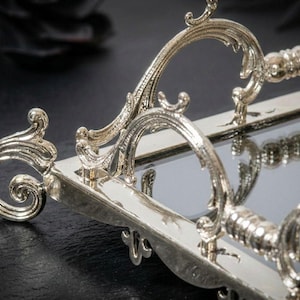 Pu&ograve; includere: Vassoio da portata rettangolare color argento con manici ornati e arricciati. Il vassoio ha una superficie a specchio e dettagli decorativi sui bordi. Le maniglie presentano un design curvo con intagli intricati.