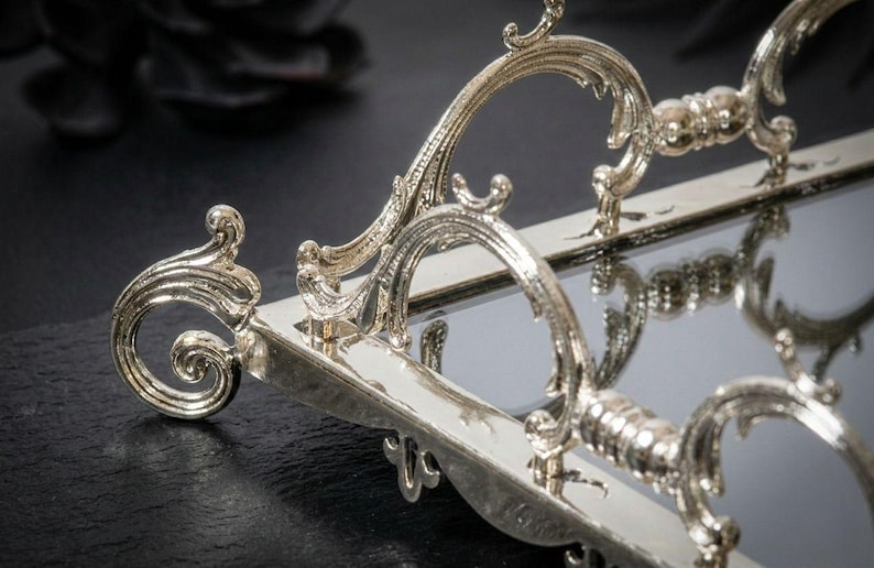 Pu&ograve; includere: Vassoio da portata rettangolare color argento con manici ornati e arricciati. Il vassoio ha una superficie a specchio e dettagli decorativi sui bordi. Le maniglie presentano un design curvo con intagli intricati.