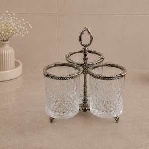 May include: A decorative bathroom organizer with two clear, textured glass cups set in a bronze-colored metal frame. The frame has an ornate design with a handle at the top. The cups are surrounded by a decorative metal trim.