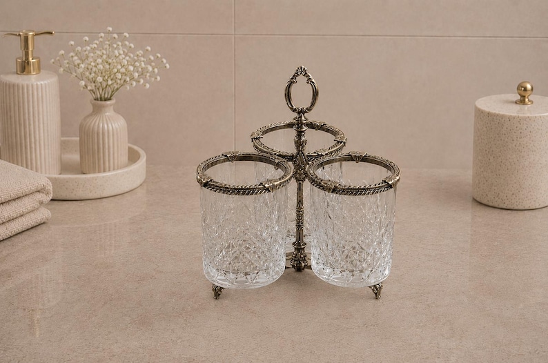 May include: A decorative bathroom organizer with two clear, textured glass cups set in a bronze-colored metal frame. The frame has an ornate design with a handle at the top. The cups are surrounded by a decorative metal trim.