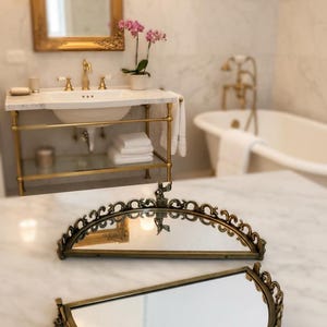 May include: Two antique brass-toned vanity mirrors with ornate, curved frames. The mirrors are displayed on a marble surface, with a bathroom vanity and bathtub in the background. The mirrors have a vintage aesthetic.