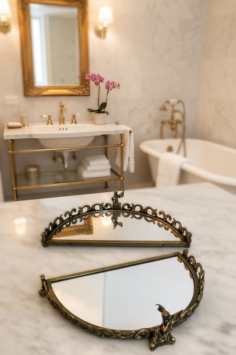 May include: Two antique brass-toned vanity mirrors with ornate, curved frames. The mirrors are displayed on a marble surface, with a bathroom vanity and bathtub in the background. The mirrors have a vintage aesthetic.