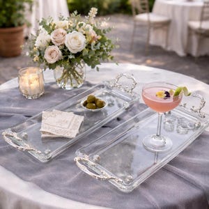 May include: Two clear glass serving trays with ornate silver handles are displayed on a gray tablecloth. One tray holds a pink cocktail garnished with a lime twist. A small bowl of olives and a stack of napkins are on the other tray. A floral arrangement and a lit candle are in the background.