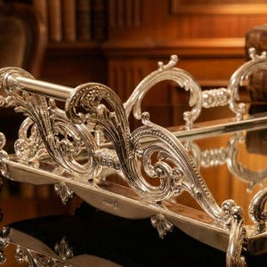 Pu&ograve; includere: Vassoio da portata argentato ornato con volute e manici intricati. Il vassoio ha una superficie a specchio ed &egrave; progettato con dettagli decorativi eleganti. Una tazza da t&egrave; e un piattino sono visibili sul vassoio, suggerendo il suo utilizzo per servire bevande.