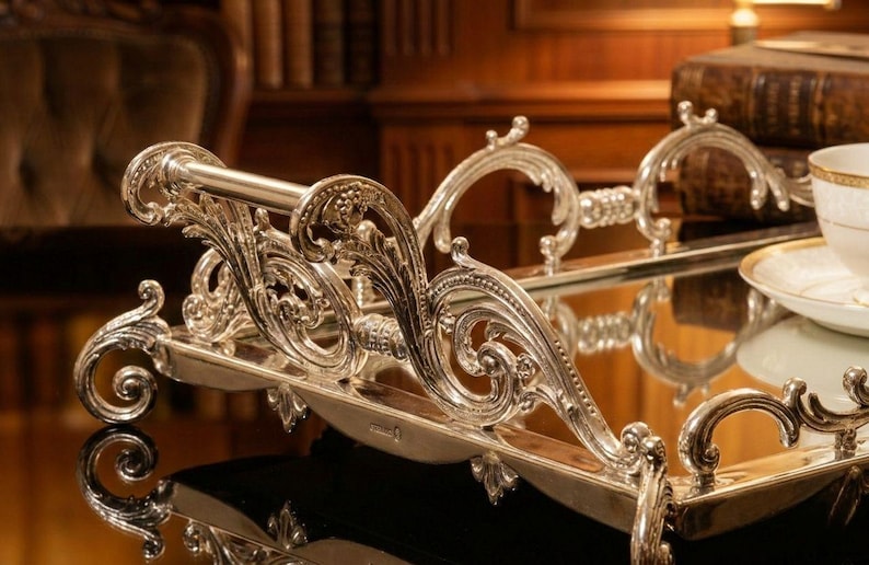 Pu&ograve; includere: Vassoio da portata argentato ornato con volute e manici intricati. Il vassoio ha una superficie a specchio ed &egrave; progettato con dettagli decorativi eleganti. Una tazza da t&egrave; e un piattino sono visibili sul vassoio, suggerendo il suo utilizzo per servire bevande.