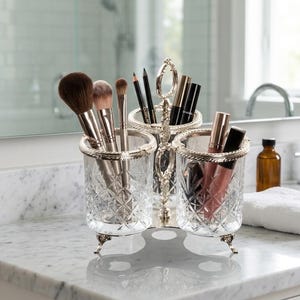 May include: A three-section makeup organizer with a silver-toned metal frame and clear, cut-glass containers. The organizer holds makeup brushes, pencils, and lip products. It sits on a marble countertop in a bathroom setting.