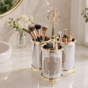 Peut inclure: Un organisateur de maquillage blanc et doré à trois compartiments, doté de récipients cylindriques avec des motifs en filigrane doré, une poignée centrale dorée et de petits pieds dorés, contenant des pinceaux et des flacons cosmétiques.