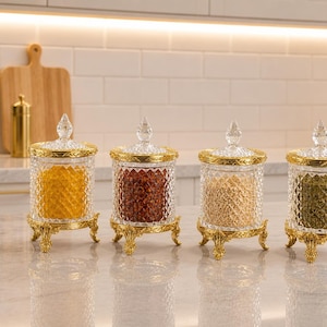 Puede incluir: Cuatro frascos de vidrio transparente con textura de rombos, cada uno con una base de metal dorado con pies ornamentados y un borde a juego. Cada frasco tiene una tapa de vidrio con un remate puntiagudo, conteniendo especias amarillas, rojas, beige y verdes.
