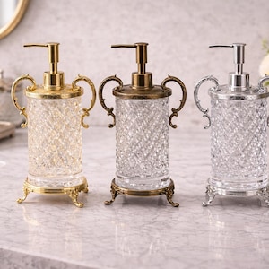 Puede incluir: Tres dispensadores de jabón decorativos con detalles ornamentados. Los dispensadores tienen un cuerpo de vidrio transparente texturizado, con detalles dorados, bronce y plateados. Cada dispensador tiene una bomba y asas decorativas. En una encimera de mármol.