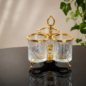 May include: A decorative three-section serving piece with a gold-colored handle and trim. The clear glass containers have a diamond pattern. The piece has three legs and is set on a reflective black surface.