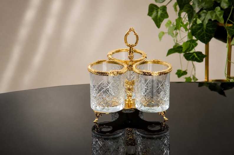 May include: A decorative three-section serving piece with a gold-colored handle and trim. The clear glass containers have a diamond pattern. The piece has three legs and is set on a reflective black surface.