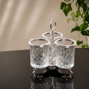 May include: A silver-toned condiment holder with three clear glass containers. Each container has a decorative diamond pattern and a silver rim. The holder has a handle and rests on three small feet. The background is neutral with a touch of greenery.