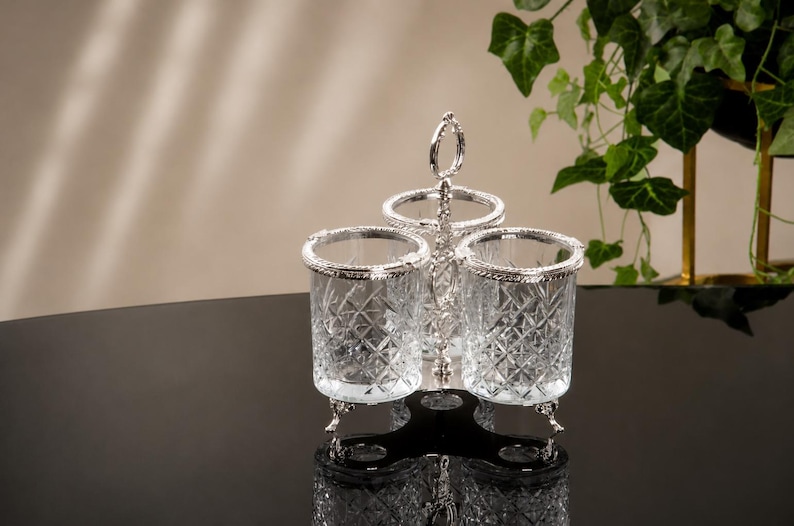 May include: A silver-toned condiment holder with three clear glass containers. Each container has a decorative diamond pattern and a silver rim. The holder has a handle and rests on three small feet. The background is neutral with a touch of greenery.
