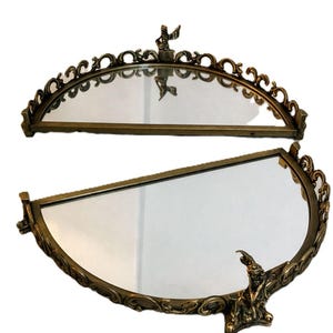 May include: Two antique, half-moon shaped mirrors with ornate, gold-toned frames. Each mirror features a decorative figure at the top and bottom. The frames have a detailed, scrollwork design, adding to their vintage aesthetic.