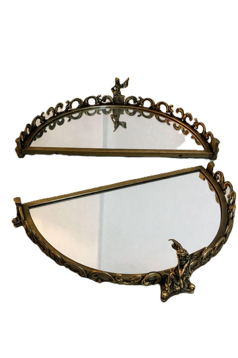 May include: Two antique, half-moon shaped mirrors with ornate, gold-toned frames. Each mirror features a decorative figure at the top and bottom. The frames have a detailed, scrollwork design, adding to their vintage aesthetic.