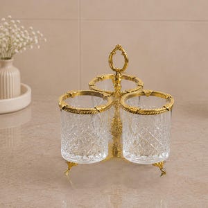 May include: A decorative three-cup holder with a gold-colored metal frame and clear, cut-glass cups. The rims and base of the holder are also gold. The holder is designed to sit on a countertop or vanity. The cups have a diamond pattern.