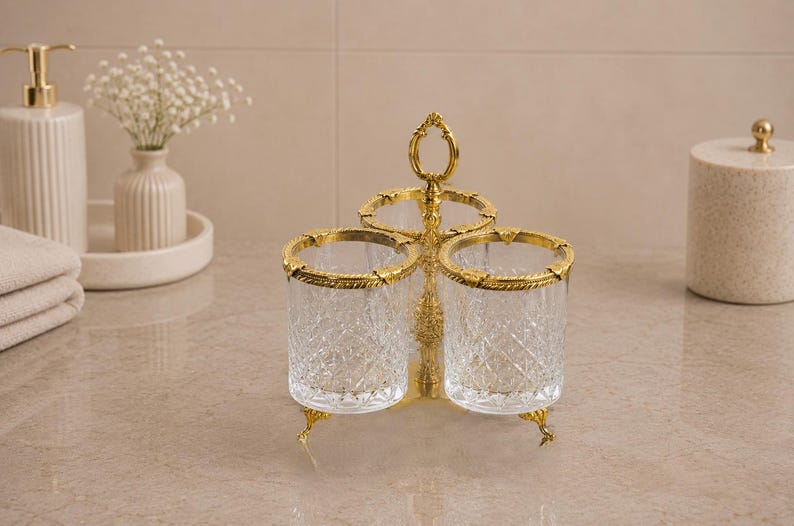 May include: A decorative three-cup holder with a gold-colored metal frame and clear, cut-glass cups. The rims and base of the holder are also gold. The holder is designed to sit on a countertop or vanity. The cups have a diamond pattern.