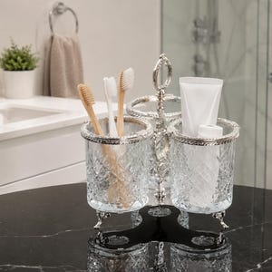 May include: A decorative bathroom organizer with three compartments. The organizer is made of clear, cut glass with silver-colored trim and a black base. It holds toothbrushes and a tube of toothpaste. The background shows a bathroom sink and shower.