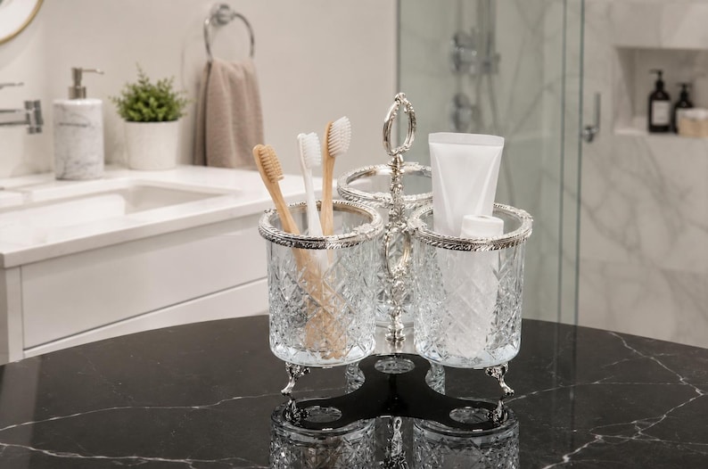 May include: A decorative bathroom organizer with three compartments. The organizer is made of clear, cut glass with silver-colored trim and a black base. It holds toothbrushes and a tube of toothpaste. The background shows a bathroom sink and shower.