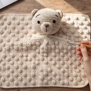 May include: A beige crochet security blanket featuring a stuffed bear head attached to a square base with a textured bobble stitch pattern. The blanket is being worked on with a wooden handled crochet hook and a length of matching yarn.