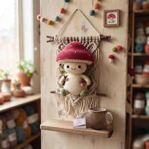 May include: A crocheted mushroom creature with a red cap and cream body, holding a small acorn, mounted on a cream macrame wall hanging. The piece features green accents, a smiling face, and is displayed above a small wooden shelf with a mug.
