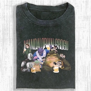 May include: Dark gray t-shirt featuring a graphic of characters from The Mandalorian and Grogu, holding mugs of beer. The text "MANDALORIAN AND GROGU" is printed across the top. The shirt is folded.