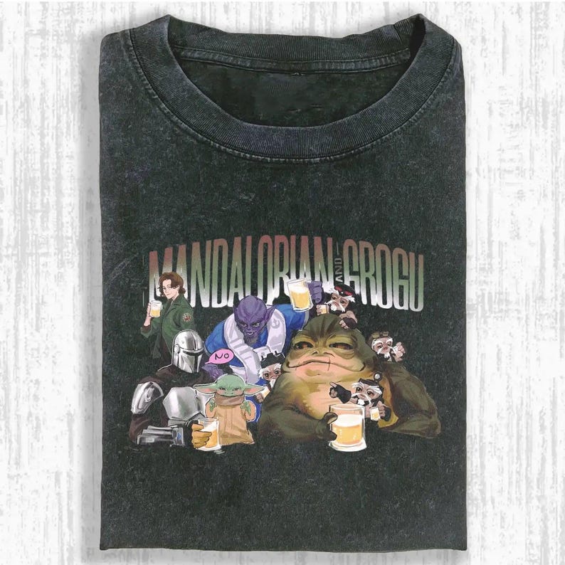 May include: Dark gray t-shirt featuring a graphic of characters from The Mandalorian and Grogu, holding mugs of beer. The text "MANDALORIAN AND GROGU" is printed across the top. The shirt is folded.